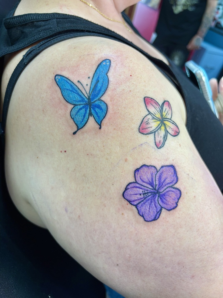 Color tattoo with blue butterfly, pink plumeria, and purple hibiscus on shoulder