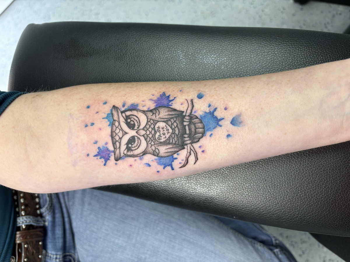 Small watercolor owl tattoo with blue and purple paint splatter accents on forearm